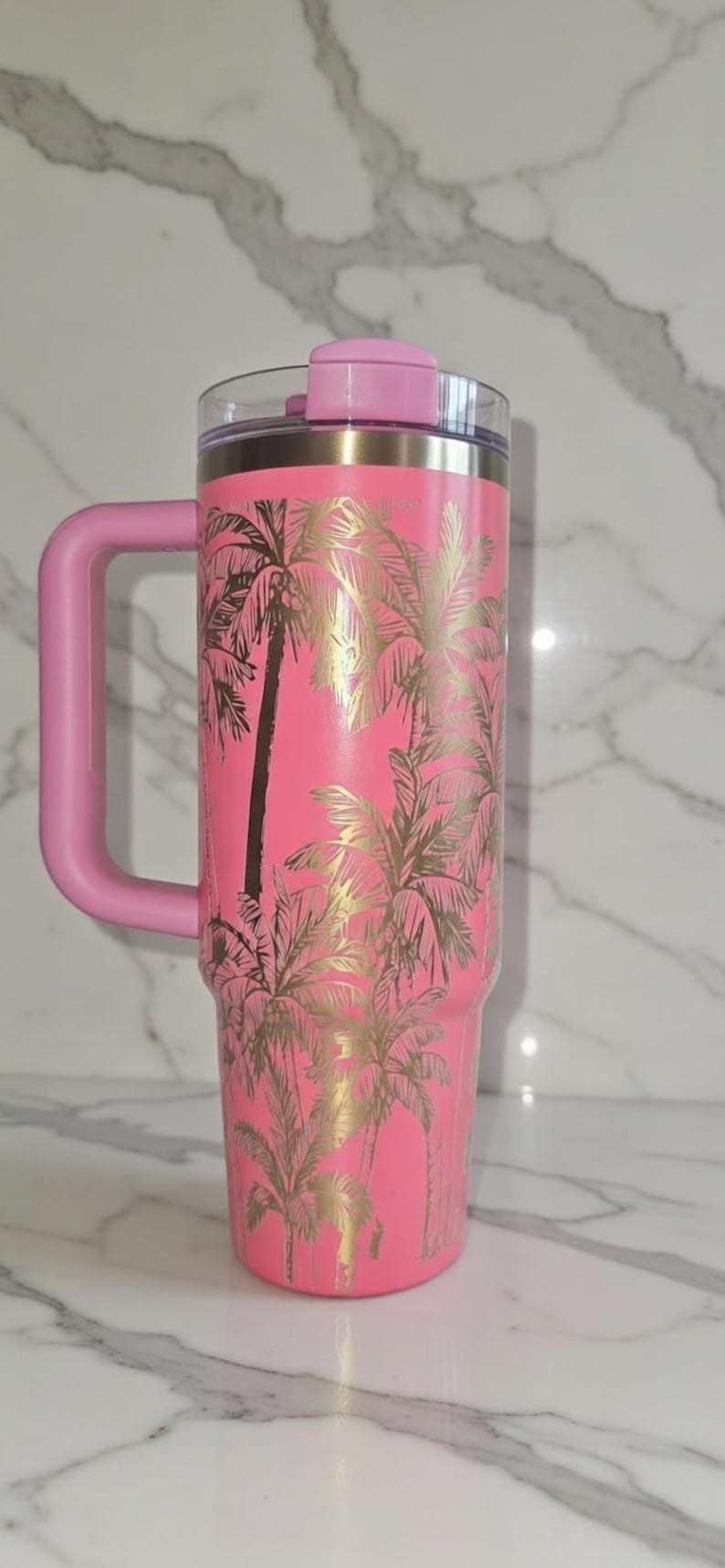 May include: Pink insulated tumbler with a handle, featuring a gold palm tree design. The tumbler has a pink lid and handle. The tumbler is approximately 10 inches tall.