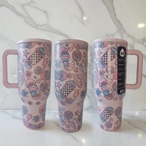 May include: Pink insulated tumbler with a handle, featuring a repeating pattern of skateboards, lightning bolts, rainbows, and flowers in pastel colors. The tumbler has a matching pink lid and handle.