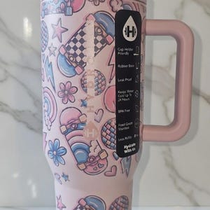 May include: A pink insulated tumbler with a handle, decorated with skateboard, star, flower, and heart graphics. The tumbler has a lid and a black label with white text. The tumbler is designed to keep water cold for up to 24 hours.