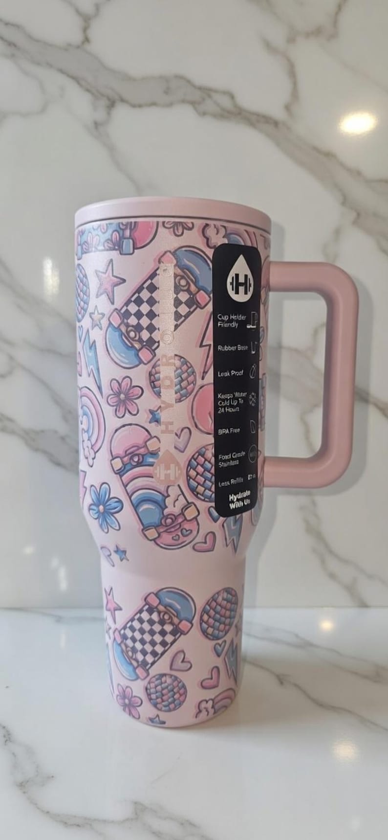 May include: A pink insulated tumbler with a handle, decorated with skateboard, star, flower, and heart graphics. The tumbler has a lid and a black label with white text. The tumbler is designed to keep water cold for up to 24 hours.