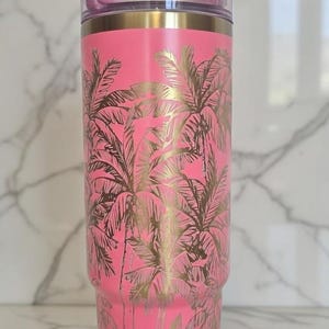 May include: A pink insulated tumbler with a gold palm tree design. The tumbler has a clear lid with a pink slider and a gold rim. The tumbler is approximately 9 inches tall.