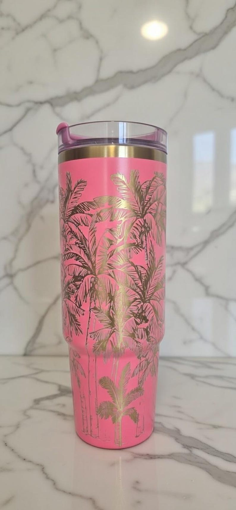 May include: A pink insulated tumbler with a gold palm tree design. The tumbler has a clear lid with a pink slider and a gold rim. The tumbler is approximately 9 inches tall.