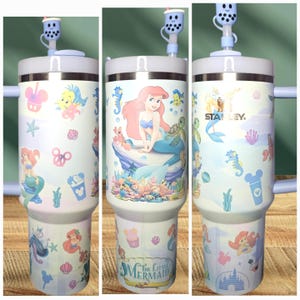 May include: A white stainless steel travel mug with a blue lid and a straw. The mug features a colorful design with Ariel from The Little Mermaid, seashells, and other Disney characters. The mug is branded with the Stanley logo.