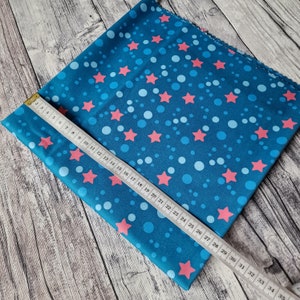 Könnte beinhalten: Ein blauer Stoff mit einem sich wiederholenden Muster aus rosa Sternen und hellblauen Kreisen. Ein Maßband liegt auf dem Stoff und zeigt eine Messung von 89 cm.