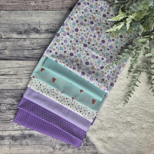 Könnte beinhalten: Ein Stapel aus sechs Stoffquadraten in verschiedenen Schattierungen von Lila und Hellblau mit unterschiedlichen Mustern. Das oberste Quadrat hat einen weißen Hintergrund mit kleinen lila und braunen Kreisen. Das zweite Quadrat hat einen hellblauen Hintergrund mit kleinen weißen Blüten und roten Erdbeeren. Das dritte Quadrat hat einen weißen Hintergrund mit kleinen braunen Herzen und roten Erdbeeren. Das vierte Quadrat ist einfarbig helllila. Das fünfte Quadrat ist einfarbig dunkellila. Das unterste Quadrat hat einen helllila Hintergrund mit kleinen weißen Punkten.