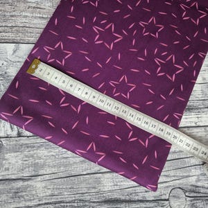 Könnte beinhalten: Ein Stück lila Stoff mit einem sich wiederholenden Muster aus rosa Sternen und Strichen. Ein weißes und gelbes Maßband liegt auf dem Stoff und zeigt Messungen bis zu 68 cm an.