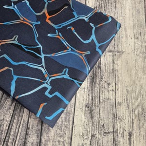 Könnte beinhalten: Gefalteter marineblauer Stoff mit einem geometrischen Muster in Blau-, Orange- und Grautönen. Das Design zeigt miteinander verbundene Linien und Formen, die eine moderne und abstrakte Ästhetik erzeugen. Der Stoff wird auf einer hellen Holzoberfläche präsentiert.