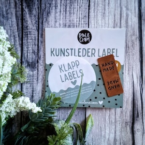 Könnte beinhalten: Nahaufnahme einer Packung Leder-Etiketten. Die Packung ist blau und weiß mit dem Text "Kunstleder Label" und "Klapp Labels" bedruckt. Ein braunes Leder-Etikett mit dem Text "Hand Made" ist an der Packung befestigt.