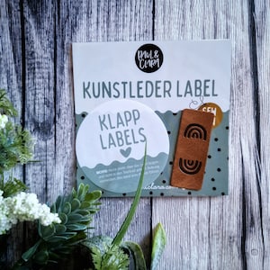 Könnte beinhalten: Nahaufnahme einer Packung brauner Leder-Etiketten mit Regenbogen-Design. Die Packung befindet sich auf einem hellen Holzuntergrund. Der Text auf der Packung lautet "Kunstleder Label" und "Klapp Labels".