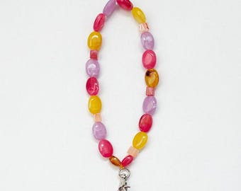 May include: A colorful phone charm with a beaded chain made of pink, yellow, and purple oval beads. The chain is attached to a silver phone ring and a black smartphone.