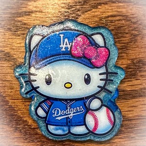 Puede incluir: Imagen de dibujos animados brillante de un personaje con uniforme y gorra de béisbol de los Dodgers, sosteniendo una pelota. La gorra muestra "LA", y el uniforme dice "Dodgers". El personaje tiene un lazo rosa brillante. Borde brillante.