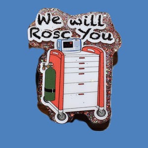 K&ouml;nnte beinhalten: Glitzernder Anstecker mit einem medizinischen Wagen mit rotem Rahmen und wei&szlig;en Schubladen, einer Sauerstoffflasche und einem Herzmonitor. Der Text "We will Rosc You" ist in schwarzer Schrift auf wei&szlig;em Hintergrund geschrieben.