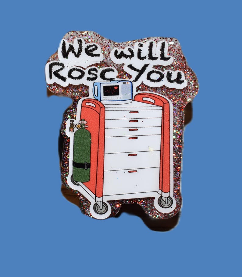 K&ouml;nnte beinhalten: Glitzernder Anstecker mit einem medizinischen Wagen mit rotem Rahmen und wei&szlig;en Schubladen, einer Sauerstoffflasche und einem Herzmonitor. Der Text "We will Rosc You" ist in schwarzer Schrift auf wei&szlig;em Hintergrund geschrieben.