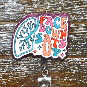 K&ouml;nnte beinhalten: Ein Ausweis-Rollhalter mit einem farbenfrohen Design. Das Abzeichen zeigt eine stilisierte Lungengrafik und die Worte "FACE DOWN SATS UP" in einer Retro-Schriftart. Das Abzeichen hat einen glitzernden lila Rand und eine einziehbare Rolle.