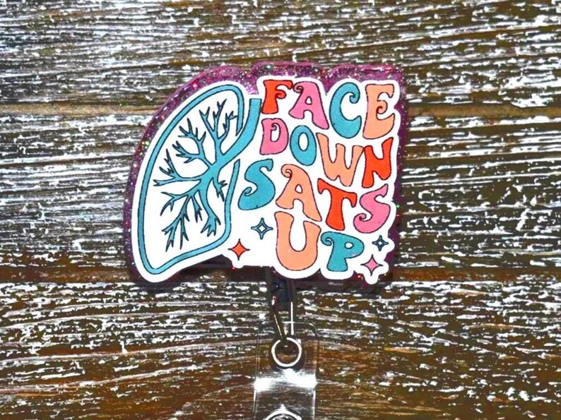 K&ouml;nnte beinhalten: Ein Ausweis-Rollhalter mit einem farbenfrohen Design. Das Abzeichen zeigt eine stilisierte Lungengrafik und die Worte "FACE DOWN SATS UP" in einer Retro-Schriftart. Das Abzeichen hat einen glitzernden lila Rand und eine einziehbare Rolle.