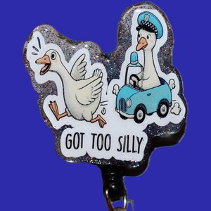 K&ouml;nnte beinhalten: Ein neuartiges einziehbares Ausweis-Jojo mit einer Cartoon-Gans, die vor einer Gans in einem blauen Auto flieht. Der Text "GOT TOO SILLY" ist auf dem Ausweis-Jojo aufgedruckt. Es hat einen silbernen Glitzerrahmen und einen schwarzen Clip.