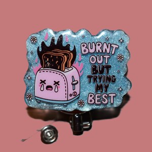 K&ouml;nnte beinhalten: Ein hellblaues, glitzerndes Ausweis-Jojo mit dem Text "BURNT OUT BUT TRYING MY BEST". Das Abzeichen zeigt einen rosa Cartoon-Toaster mit verbranntem Toast und Flammen. Das Jojo hat einen silbernen Metallclip.