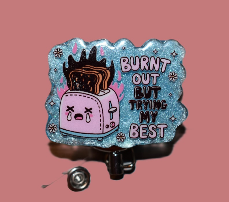 K&ouml;nnte beinhalten: Ein hellblaues, glitzerndes Ausweis-Jojo mit dem Text "BURNT OUT BUT TRYING MY BEST". Das Abzeichen zeigt einen rosa Cartoon-Toaster mit verbranntem Toast und Flammen. Das Jojo hat einen silbernen Metallclip.