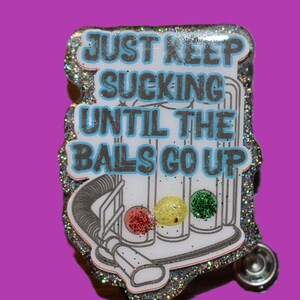 K&ouml;nnte beinhalten: Ein glitzernder Anstecker mit dem Text "JUST KEEP SUCKING UNTIL THE BALS GO UP" in Blau und Grau. Das Design beinhaltet eine Staubsauger-Grafik mit drei farbigen Kugeln: rot, gelb und gr&uuml;n. Der Anstecker hat einen silbernen Rand und einen Nadelverschluss.