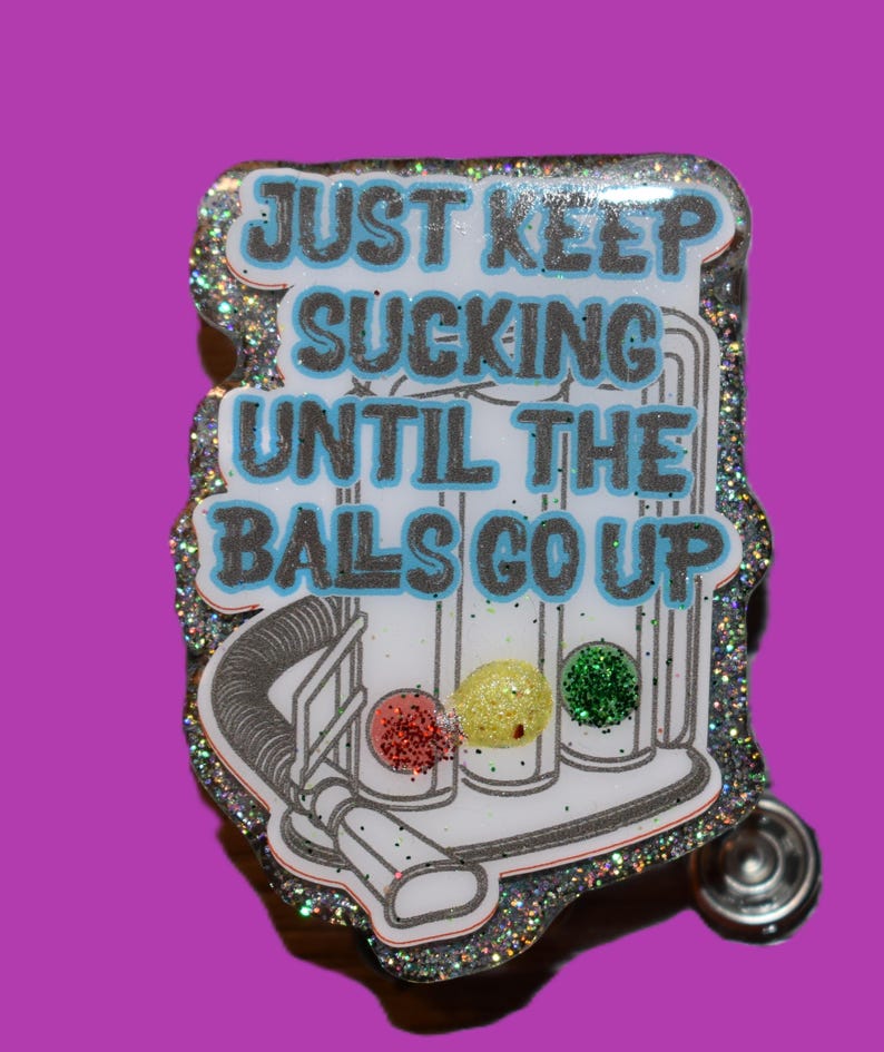 K&ouml;nnte beinhalten: Ein glitzernder Anstecker mit dem Text "JUST KEEP SUCKING UNTIL THE BALS GO UP" in Blau und Grau. Das Design beinhaltet eine Staubsauger-Grafik mit drei farbigen Kugeln: rot, gelb und gr&uuml;n. Der Anstecker hat einen silbernen Rand und einen Nadelverschluss.
