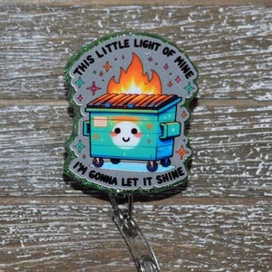 K&ouml;nnte beinhalten: Ein Ausweis-Rollhalter mit einem Design einer t&uuml;rkisfarbenen M&uuml;lltonne, Flammen und dem Text "THIS LITTLE LIGHT OF MINE I'M GONNA LET IT SHINE". Der Rollhalter hat einen transparenten Kunststoffclip und ein einziehbares Kabel.