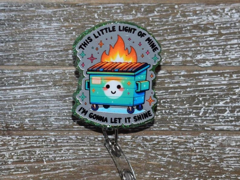 K&ouml;nnte beinhalten: Ein Ausweis-Rollhalter mit einem Design einer t&uuml;rkisfarbenen M&uuml;lltonne, Flammen und dem Text "THIS LITTLE LIGHT OF MINE I'M GONNA LET IT SHINE". Der Rollhalter hat einen transparenten Kunststoffclip und ein einziehbares Kabel.