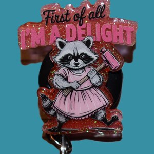 K&ouml;nnte beinhalten: Ein einziehbarer Ausweis-Clip mit einem Waschb&auml;ren in einem rosa Kleid, der einen rosa Hammer h&auml;lt. Der Text "First of all I'M A DELIGHT" steht &uuml;ber dem Waschb&auml;ren. Die Rolle hat einen glitzernden roten Hintergrund und eine schwarze R&uuml;ckseite.