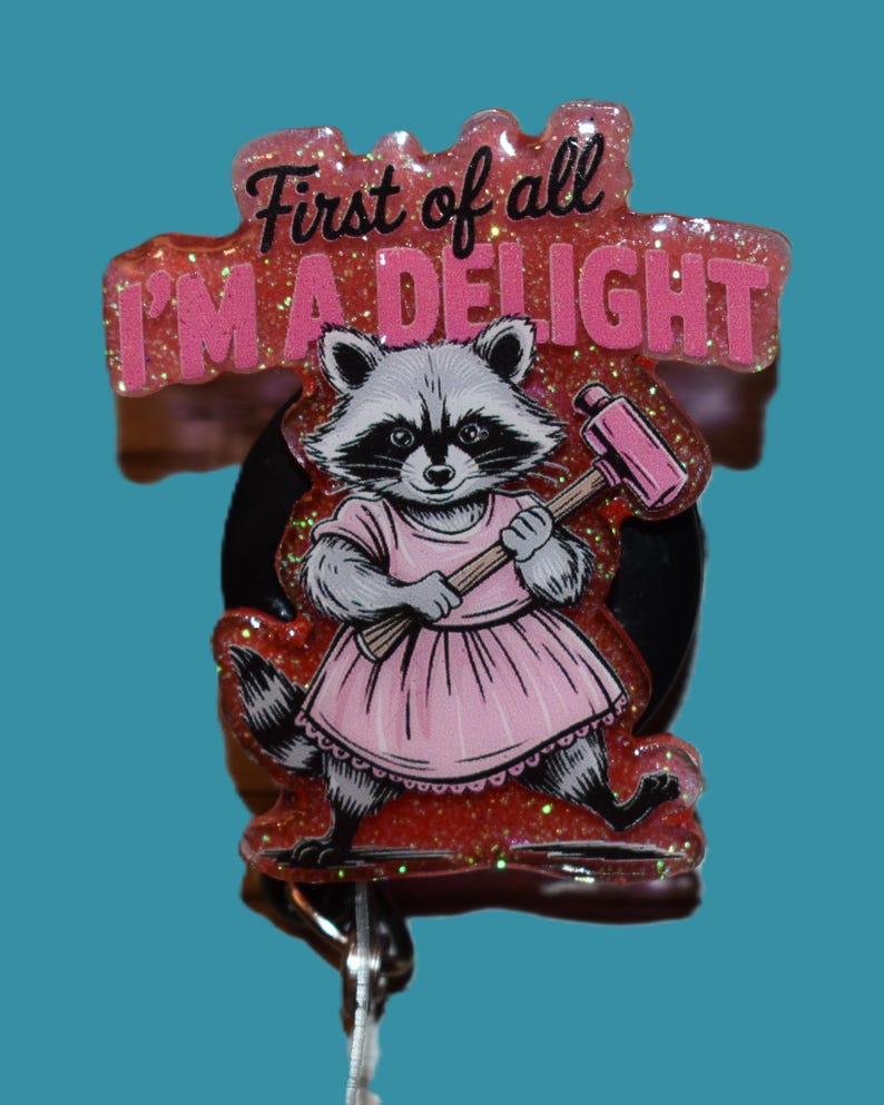 K&ouml;nnte beinhalten: Ein einziehbarer Ausweis-Clip mit einem Waschb&auml;ren in einem rosa Kleid, der einen rosa Hammer h&auml;lt. Der Text "First of all I'M A DELIGHT" steht &uuml;ber dem Waschb&auml;ren. Die Rolle hat einen glitzernden roten Hintergrund und eine schwarze R&uuml;ckseite.