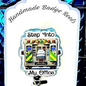Può includere: Un portabadge con un design di ambulanza. L'immagine include il testo "Step Into My Office". Il portabadge è bianco con un bordo smerlato e le parole "Handmade Badge Reels" in corsivo.
