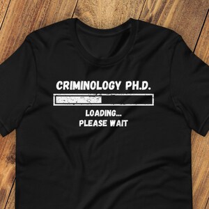 May include: Black t-shirt with white text that reads "Criminology Ph.D. Loading... Please Wait".