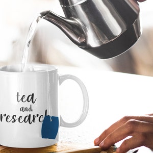 May include: White ceramic mug with the text "tea and research" and a blue tea bag hanging from the rim. Hot water is being poured into the mug from a silver teapot.