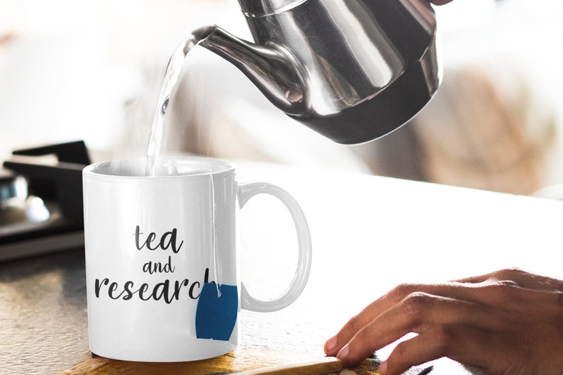 May include: White ceramic mug with the text "tea and research" and a blue tea bag hanging from the rim. Hot water is being poured into the mug from a silver teapot.
