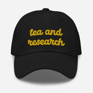 Op de afbeelding: Zwarte baseballpet met de tekst "tea and research" geborduurd in geel.