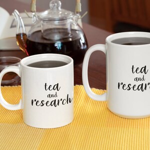 May include: Two white ceramic mugs with black text that reads "tea and research". The mugs are filled with a dark liquid and are sitting on a yellow placemat.
