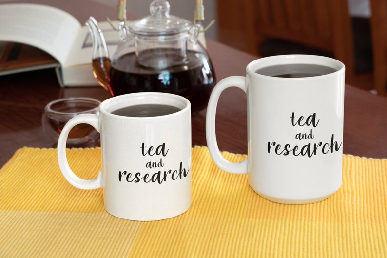 May include: Two white ceramic mugs with black text that reads "tea and research". The mugs are filled with a dark liquid and are sitting on a yellow placemat.