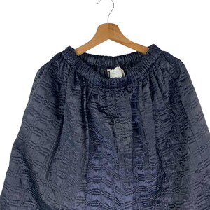 May include: A navy blue skirt with a textured, quilted pattern. The skirt is hanging on a wooden hanger against a white background. The fabric appears to be a soft, flowing material.