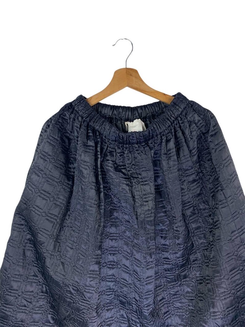 May include: A navy blue skirt with a textured, quilted pattern. The skirt is hanging on a wooden hanger against a white background. The fabric appears to be a soft, flowing material.