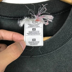 Puede incluir: Camiseta negra con una etiqueta blanca que dice "100% algod&oacute;n preencogido Ensamblado en El Salvador de componentes de EE. UU. Cuidado en el reverso" y una etiqueta de talla que dice "M".