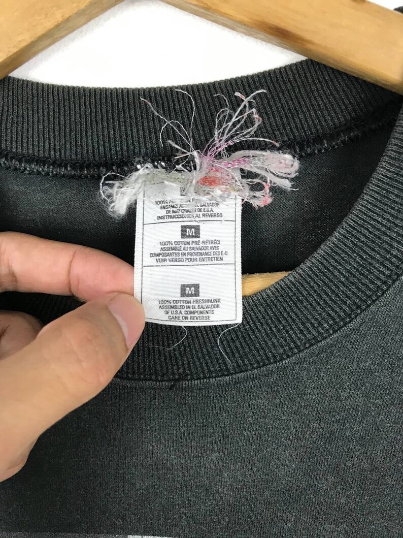 Puede incluir: Camiseta negra con una etiqueta blanca que dice "100% algod&oacute;n preencogido Ensamblado en El Salvador de componentes de EE. UU. Cuidado en el reverso" y una etiqueta de talla que dice "M".