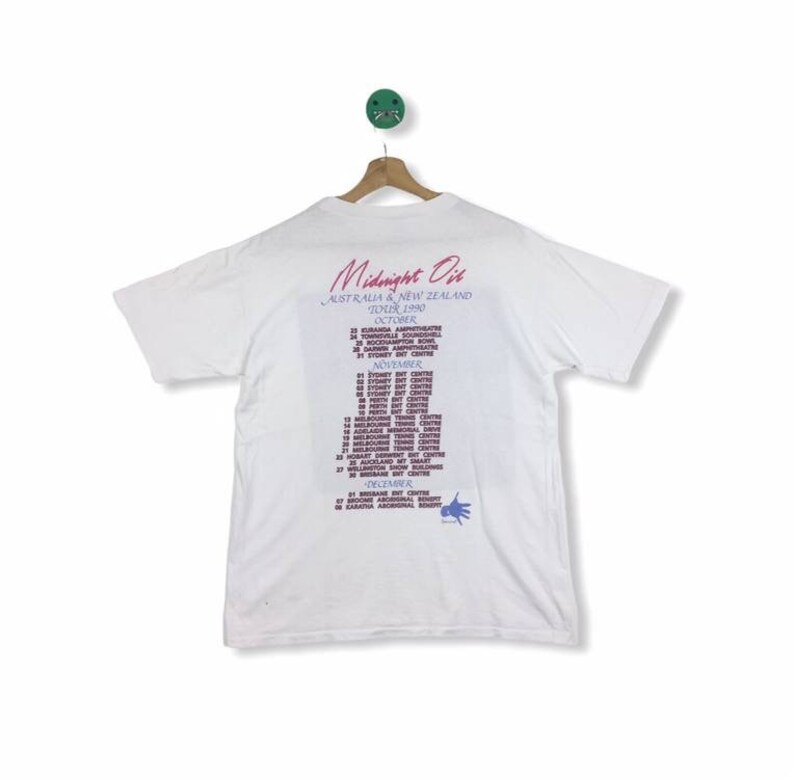 May include: White t-shirt with a Midnight Oil tour poster printed on the back. The poster lists the dates and locations for the Australian and New Zealand tour in 1990. The tour dates are in October, November, and December.