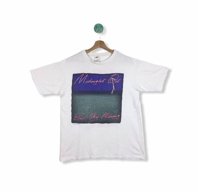 May include: White t-shirt with a graphic of the band Midnight Oil's album cover for "Blue Sky Mining". The album cover features a blue and purple sky with a lightning bolt and the text "Midnight Oil" in pink.