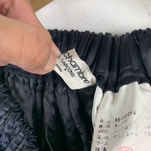 May include: A close-up shot of dark blue fabric with a gathered texture. A white label with the text "chambre des garcons" is visible, alongside a smaller tag with Japanese text. The fabric is held by a hand.