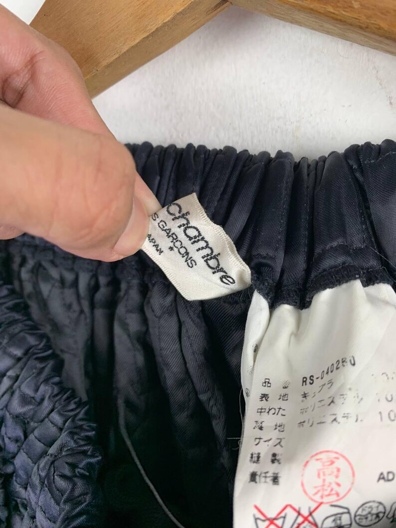 May include: A close-up shot of dark blue fabric with a gathered texture. A white label with the text "chambre des garcons" is visible, alongside a smaller tag with Japanese text. The fabric is held by a hand.