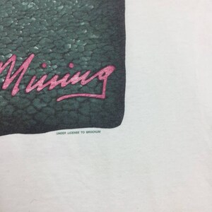 May include: A white t-shirt with a green and black graphic of a crocodile with the word "Mining" written in pink lettering. The text "Under License to Brockum" is printed in black below the graphic.