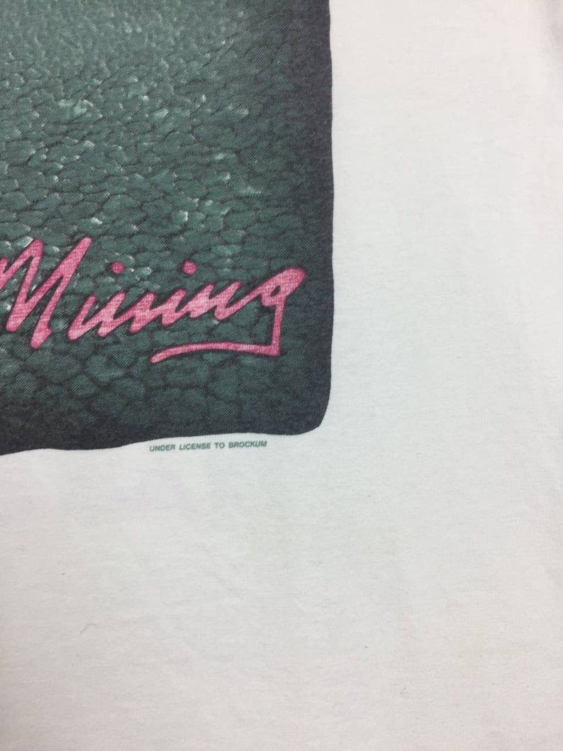 May include: A white t-shirt with a green and black graphic of a crocodile with the word "Mining" written in pink lettering. The text "Under License to Brockum" is printed in black below the graphic.