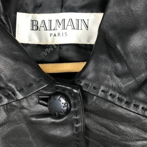 May include: Black leather jacket with a Balmain Paris label and a button closure. The jacket has a collar and a stitched design.