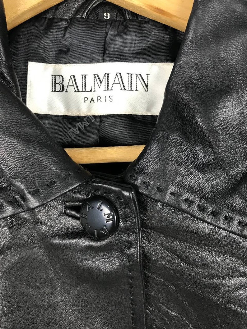 May include: Black leather jacket with a Balmain Paris label and a button closure. The jacket has a collar and a stitched design.