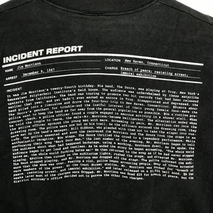 Puede incluir: Texto blanco y negro sobre una camiseta negra. El texto dice "Informe de incidente" e incluye detalles sobre el arresto de Jim Morrison el 9 de diciembre de 1967 en New Haven, Connecticut. Los cargos fueron alteraci&oacute;n del orden p&uacute;blico, resistencia al arresto y exhibici&oacute;n inmoral.