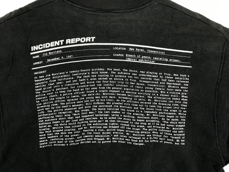 Puede incluir: Texto blanco y negro sobre una camiseta negra. El texto dice "Informe de incidente" e incluye detalles sobre el arresto de Jim Morrison el 9 de diciembre de 1967 en New Haven, Connecticut. Los cargos fueron alteraci&oacute;n del orden p&uacute;blico, resistencia al arresto y exhibici&oacute;n inmoral.
