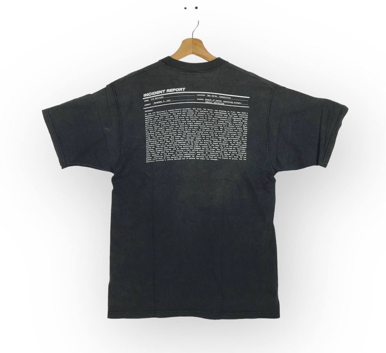 Puede incluir: Camiseta negra con una impresi&oacute;n blanca en la espalda que dice "INCIDENT REPORT" e incluye un bloque de texto. El texto incluye las palabras "Asunto: El evento", "Fecha: 8 de junio de 1997" y "Ubicaci&oacute;n: La parte de atr&aacute;s de la pizzer&iacute;a de John".
